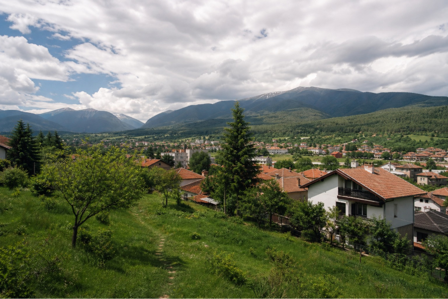 Панорамна гледка към планински град с къщи, зелени поляни и високи планински върхове.