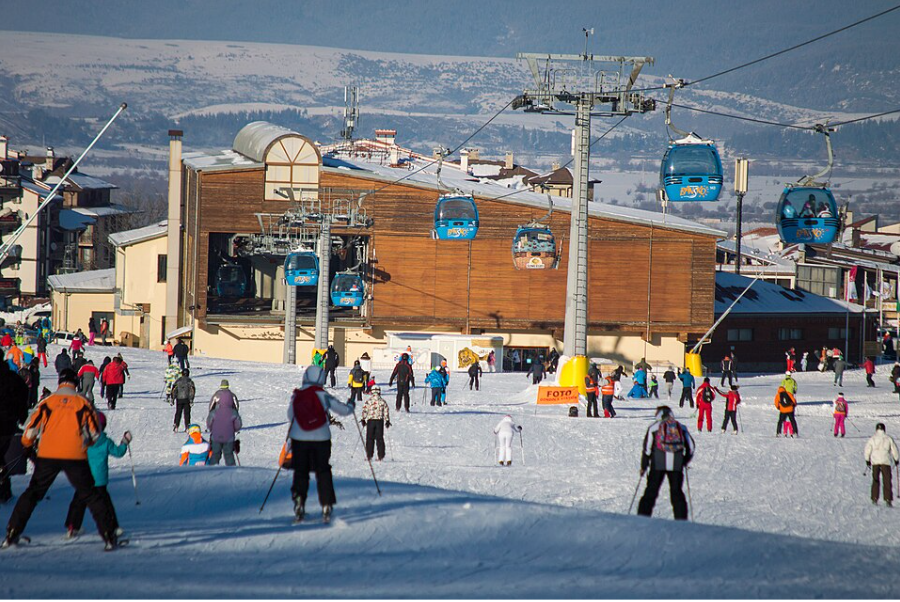 Синя кабина от лифт с надпис „Bansko“ пристига на станцията на гондолата. Под нея има ски писта със скиори, а отзад се виждат заснежени върхове.